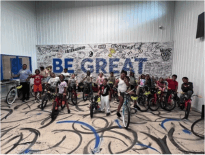 Participants from the Learn and Earn a Bike 2025 program lined up qith their bikes in fornt of am ural that reads "BE GREAT"